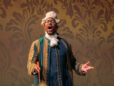 Singer in renaissance clothes and a wig singing in front of an ornate wallpaper projection